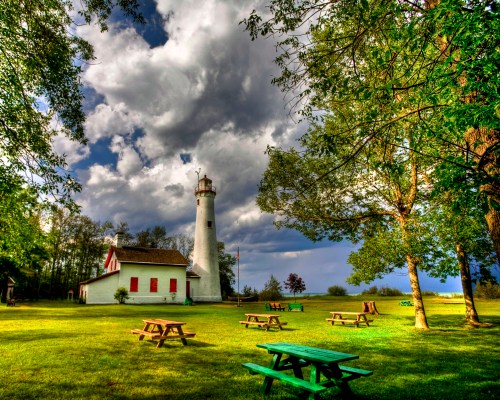 A scenic lighthouse surrounded by trees and picnic tables under a cloudy sky.