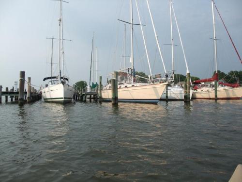 A calm marina scene with several sailboats docked along wooden piers, reflecting in the water under a cloudy sky.