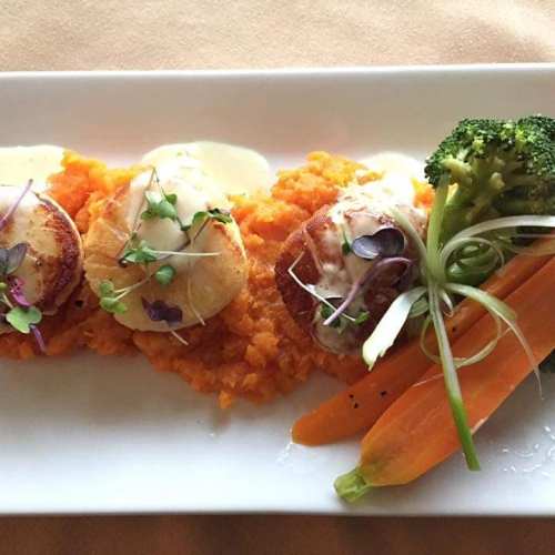 A gourmet dish featuring three scallops on a bed of sweet potato puree, garnished with broccoli and carrots.