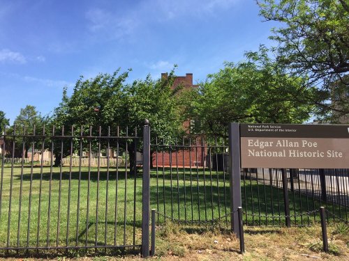 Sign for the Edgar Allan Poe National Historic Site, surrounded by greenery and a fence, with a historic building in the background.