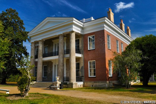 A historic mansion with columns, red brick, and a blue sky, surrounded by trees and a gravel pathway.