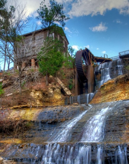 A historic watermill beside a cascading waterfall, surrounded by trees and a blue sky with scattered clouds.