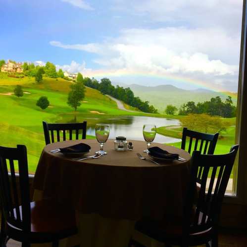 A table set for dining with glasses and silverware, overlooking a scenic view of a golf course and rainbow.