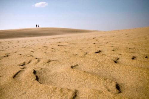 Two silhouettes stand on a sandy dune under a clear blue sky, with footprints leading towards them.