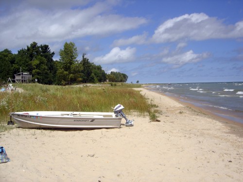 A small boat rests on a sandy beach beside a calm lake, with trees and a cloudy sky in the background.