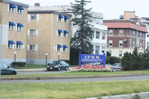 Sign for Lynn, Massachusetts, with nearby residential buildings and landscaped flowers in the foreground.