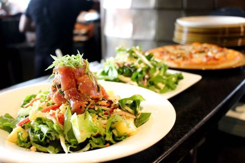 A vibrant salad topped with tuna and greens, with another salad and a pizza in the background.