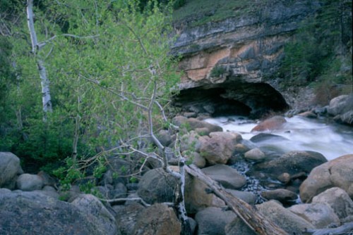 A serene river flows past large rocks and greenery, leading to a cave in a rocky cliff.