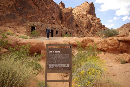 A person walks towards stone cabins in a desert landscape, with rocky formations and wildflowers nearby.