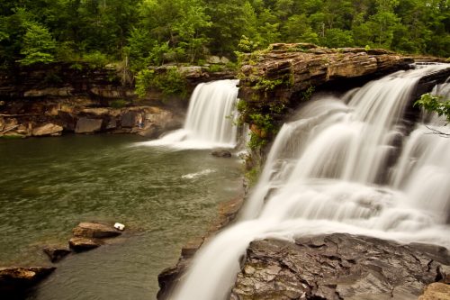 A serene waterfall cascades over rocky cliffs into a tranquil pool, surrounded by lush green trees.