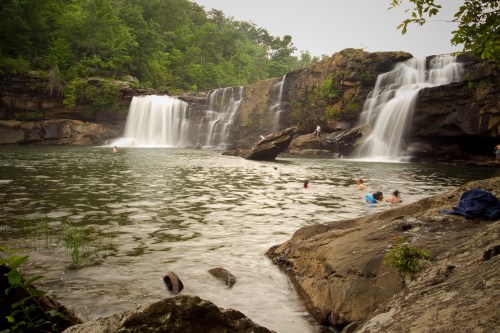 A serene waterfall cascades into a calm pool, with people swimming and lush greenery surrounding the scene.