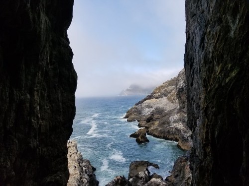 View of a rocky coastline through a narrow opening, with waves crashing and mist over the distant hills.