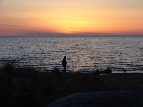 Silhouette of a person walking along the shore at sunset, with calm waters and a colorful sky.