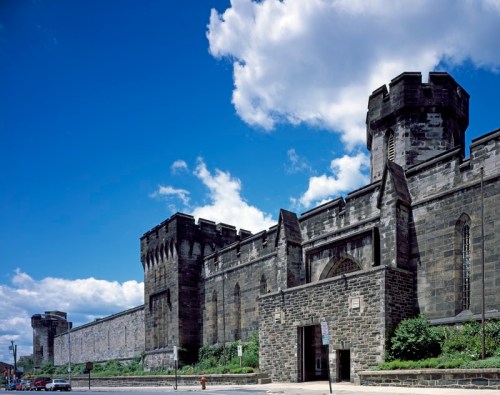 Historic stone prison building with towers under a blue sky and scattered clouds. Greenery at the base.