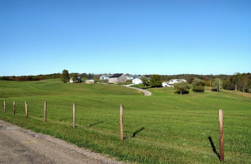 A scenic rural landscape with green fields, a winding road, and farm buildings under a clear blue sky.