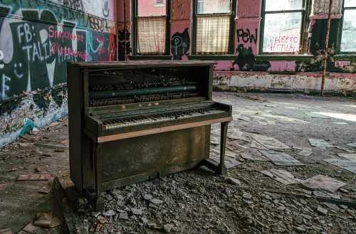 An old, dusty piano sits in a dilapidated room with graffiti-covered walls and debris scattered on the floor.