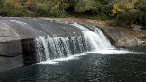 A serene waterfall cascading over smooth rocks into a calm pool, surrounded by lush greenery and autumn foliage.