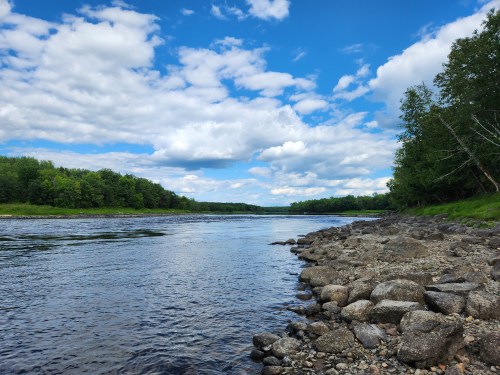 A serene river scene with rocky banks, lush green trees, and a blue sky filled with fluffy clouds.