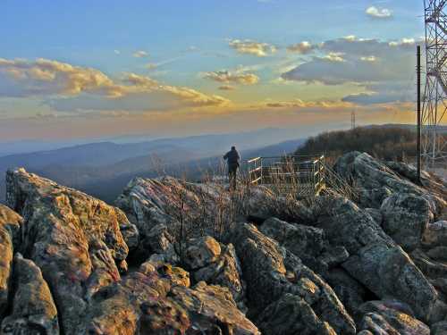 A person stands on rocky terrain at sunset, overlooking rolling hills and a colorful sky with clouds.