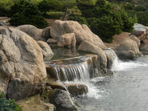 A serene scene of a small waterfall cascading over rocks into a calm body of water, surrounded by greenery.