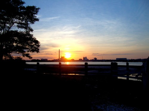 Sunset over a calm body of water, with silhouettes of trees and buildings against a colorful sky.