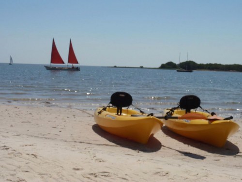 Two yellow kayaks on a sandy beach with a calm sea and sailboats in the background under a clear blue sky.