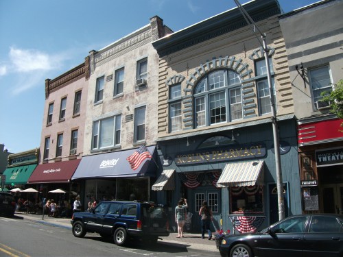 Historic storefronts line a street, featuring a pharmacy and outdoor dining, with people enjoying the day.