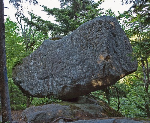 A large, balanced boulder sits atop a smaller rock, surrounded by lush green trees in a forested area.