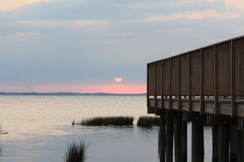 A serene sunset over calm waters, with a wooden pier extending into the scene and grass visible in the foreground.
