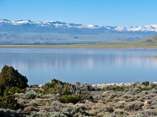 A serene landscape featuring a calm lake, green hills, and snow-capped mountains under a clear blue sky.