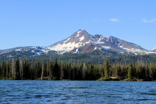 A scenic view of a snow-capped mountain reflecting in a calm lake, surrounded by lush green forests under a clear blue sky.