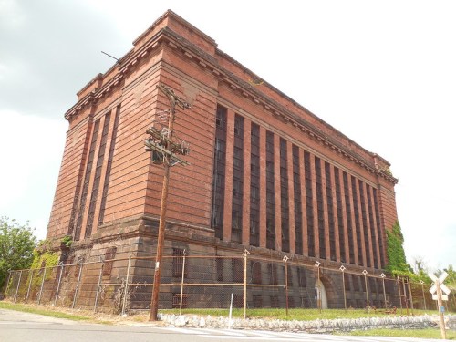 A large, abandoned brick building with tall windows, surrounded by a chain-link fence and overgrown vegetation.