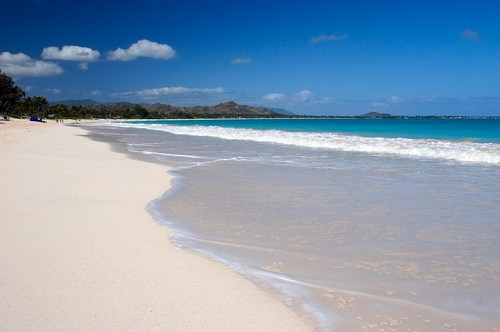 A serene beach with soft white sand, gentle waves, and a clear blue sky, framed by distant mountains.
