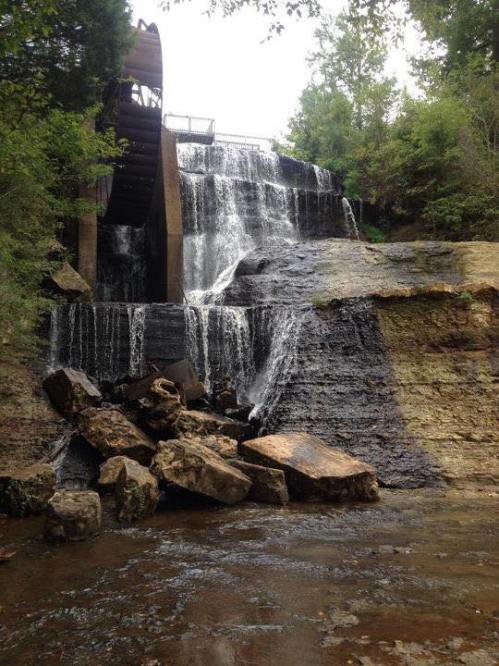 A cascading waterfall flows over rocky terrain, surrounded by lush greenery and a wooden structure above.