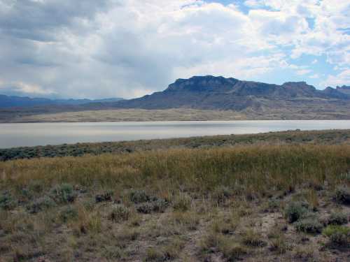 A serene landscape featuring a lake surrounded by grassy fields and distant mountains under a cloudy sky.