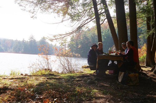 A group of four people enjoying a meal at a lakeside picnic table surrounded by trees in autumn.