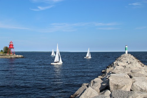 Sailboats glide on calm waters near a rocky pier, with a red lighthouse and a green marker under a clear blue sky.