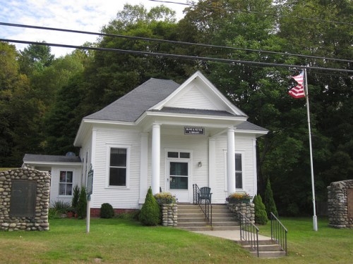 A white building with a porch, surrounded by greenery, featuring an American flag and stone pillars at the entrance.