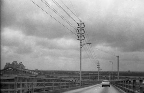 A black and white photo of a road leading over a bridge, with power lines and a cloudy sky in the background.