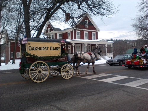 A horse-drawn wagon labeled "Oakhurst Dairy" passes by a red house in a snowy setting during a parade.