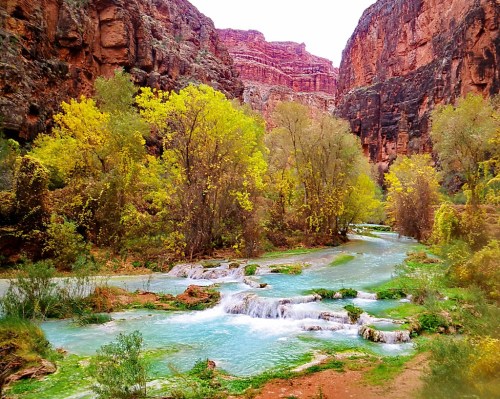 A serene river flows through a canyon, surrounded by vibrant green trees and rocky cliffs under a cloudy sky.
