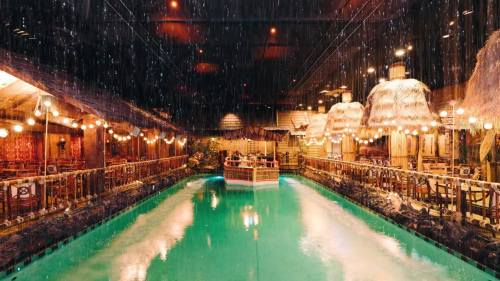 A tropical-themed restaurant with a pool, thatched roofs, and decorative lights, creating a vibrant, rainy atmosphere.