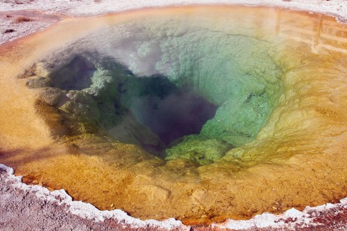 A vibrant geothermal hot spring with clear blue water surrounded by yellow and orange mineral deposits.
