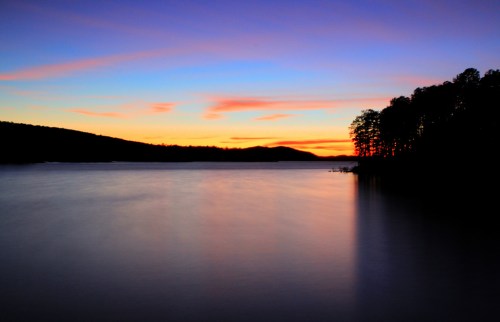 A serene lake at sunset, with vibrant colors in the sky and silhouettes of trees along the shore.
