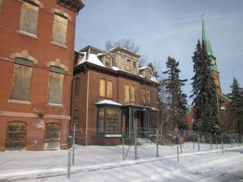 Abandoned brick building with boarded windows, surrounded by snow and a fence, near a church with a green steeple.