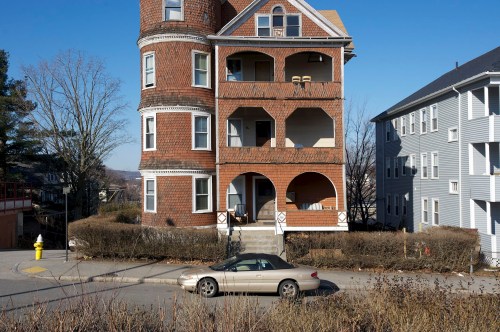 A three-story brick building with arched balconies, surrounded by shrubs and a parked car on a quiet street.