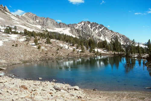 A serene mountain landscape featuring a clear lake, rocky shore, and snow-capped peaks under a blue sky.