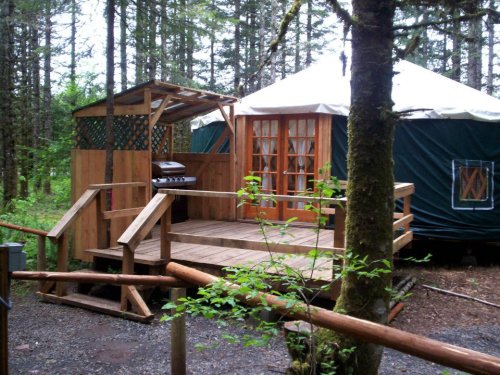 A wooden deck with a grill leads to a yurt surrounded by trees in a forested area.