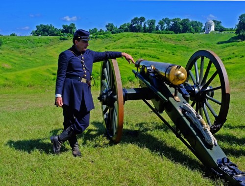 A soldier in historical uniform leans against a cannon on a grassy field with rolling hills in the background.