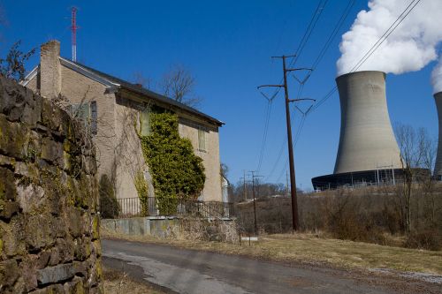 A rural scene featuring a house with ivy, power lines, and cooling towers emitting steam in the background.